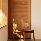 The Penguin Wooden Lounge Chair in natural oak, positioned in a sunlit room against a matching wooden door.