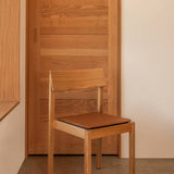 Mauro Dining Chair in natural oak with a brown leather seat cushion, positioned against a wooden door and light wall.
