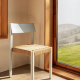 Mauro Dining Chair in Linen Blue Oak with a woven paper cord seat, standing in a sunlit corner by a window.