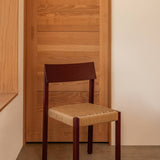Mauro Dining Chair in French red oak with a handwoven paper cord seat, set against a minimalist wooden door.